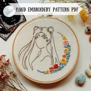 May include: A hand embroidery pattern featuring a line drawing of a woman with long hair and a crescent moon with floral accents. The pattern is displayed in a wooden embroidery hoop.