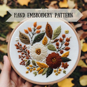 Könnte beinhalten: Handstickmuster mit einem floralen Design mit weißen, orangefarbenen, gelben und grünen Blumen und Blättern. Das Muster ist auf einem weißen Stoff gestickt und in einem Holzreifen gerahmt.