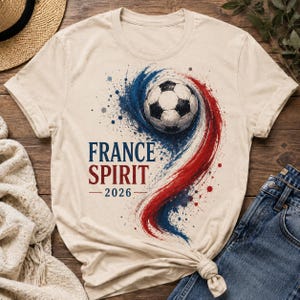May include: Beige t-shirt featuring a soccer ball design with blue and red paint strokes. The text "FRANCE SPIRIT 2026" is printed in blue and red. The shirt is made of a soft material.