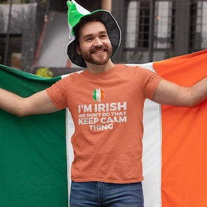 May include: An orange t-shirt with the text "I'M IRISH WE DON'T DO THAT KEEP CALM THING" and an Irish flag emblem. The person is wearing a green and white hat and holding an Irish flag. The flag is green, white, and orange.