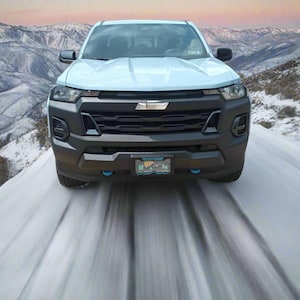 Puede incluir: Una camioneta azul claro conduciendo por una carretera de montaña nevada. La camioneta tiene una parrilla y parachoques negros, con una placa visible. El fondo presenta montañas cubiertas de nieve y una puesta de sol.