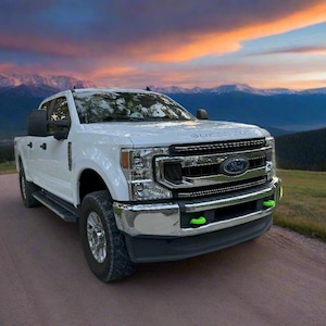 以下が含まれることがあります： 白いフォード・スーパーデューティのピックアップトラックが未舗装の道路に駐車されています。トラックはクロムのアクセント、黒いサイドミラー、フロントバンパーの緑色のアクセントが特徴です。背景には山脈とカラフルな夕焼けが写っています。