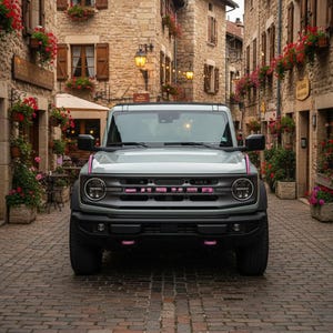 Puede incluir: Un SUV gris claro con marco cuadrado, faros redondos y una parrilla gris oscuro con letras rosas. El vehículo tiene espejos laterales negros, parachoques negros y detalles rosas en el capó y los ganchos de remolque delanteros sobre una calle adoquinada.