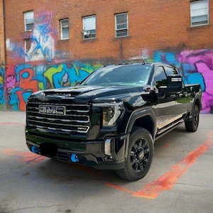 以下が含まれることがあります： カラフルなグラフィティアートが施されたレンガ造りの建物の前に駐車された黒いGMCトラック。トラックは黒いホイールとフロントバンパーに青いアクセントが付いています。GMCのロゴがフロントグリルに表示されています。