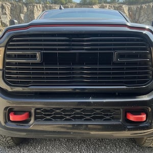 May include: Front view of a black pickup truck with a black grille and red accents. The truck has large headlights and a prominent front bumper with red tow hooks. The background shows a rocky terrain.