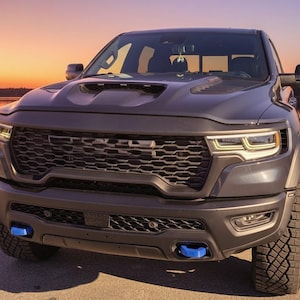 May include: A dark gray pickup truck with a black grill and blue accents. The truck is parked on a paved surface near a body of water. The sky is a gradient of orange and yellow.