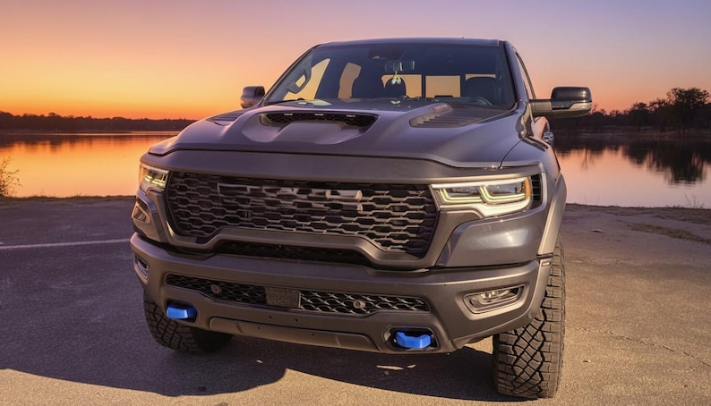 May include: A dark gray pickup truck with a black grill and blue accents. The truck is parked on a paved surface near a body of water. The sky is a gradient of orange and yellow.