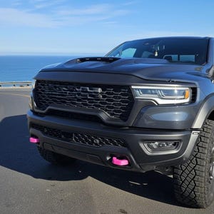 May include: A dark gray pickup truck with a black grille and pink accents on the front bumper. The truck is parked on a winding road with the ocean in the background. The tires are large and off-road ready.
