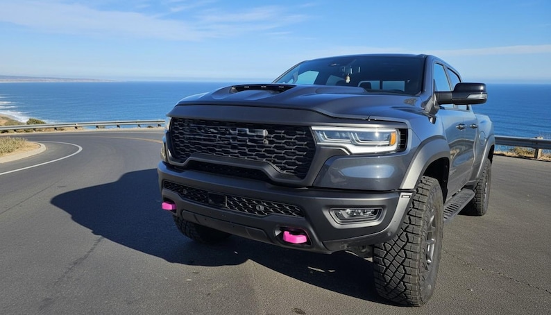May include: A dark gray pickup truck with a black grille and pink accents on the front bumper. The truck is parked on a winding road with the ocean in the background. The tires are large and off-road ready.