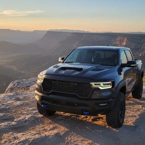 May include: A dark blue pickup truck with black accents is parked on a rocky cliff overlooking a canyon at sunset. The truck has a black grille, hood scoop, and blue tow hooks. The landscape features layered rock formations.