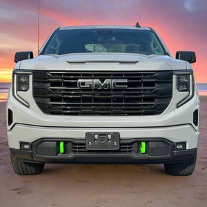 Puede incluir: Camioneta GMC blanca con una parrilla negra y el logotipo de GMC. La camioneta está estacionada en una playa con una puesta de sol en el fondo.