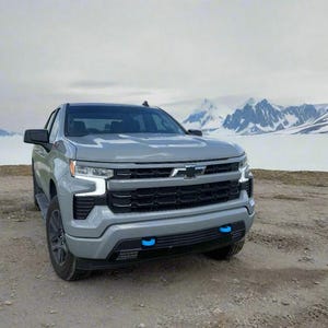May include: A gray Chevrolet Silverado pickup truck is parked on a dirt road with a snowy mountain range in the background. The truck has black accents, blue tow hooks, and a black grille.