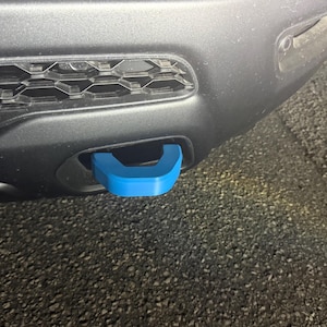 May include: A close-up of a vehicle's front bumper featuring a bright blue tow hook. The hook is U-shaped and contrasts with the dark gray bumper and the black grill. The image is taken outdoors.