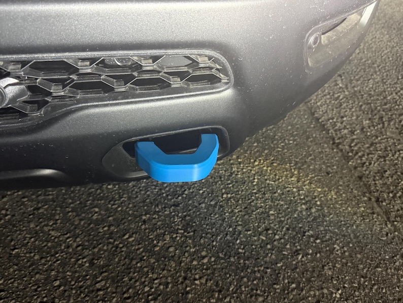 May include: A close-up of a vehicle's front bumper featuring a bright blue tow hook. The hook is U-shaped and contrasts with the dark gray bumper and the black grill. The image is taken outdoors.