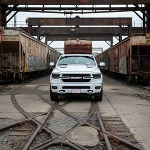 May include: A white pickup truck is centered on railroad tracks, flanked by two freight train cars. The truck has a black grill and headlights. The scene is set in an industrial area with rusty metal structures overhead.