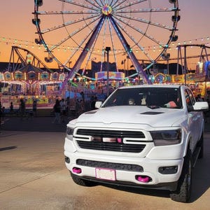 May include: A white pickup truck with pink accents is parked in front of a brightly lit Ferris wheel and carnival rides at dusk. The sky transitions from orange to purple. The truck has a pink license plate.