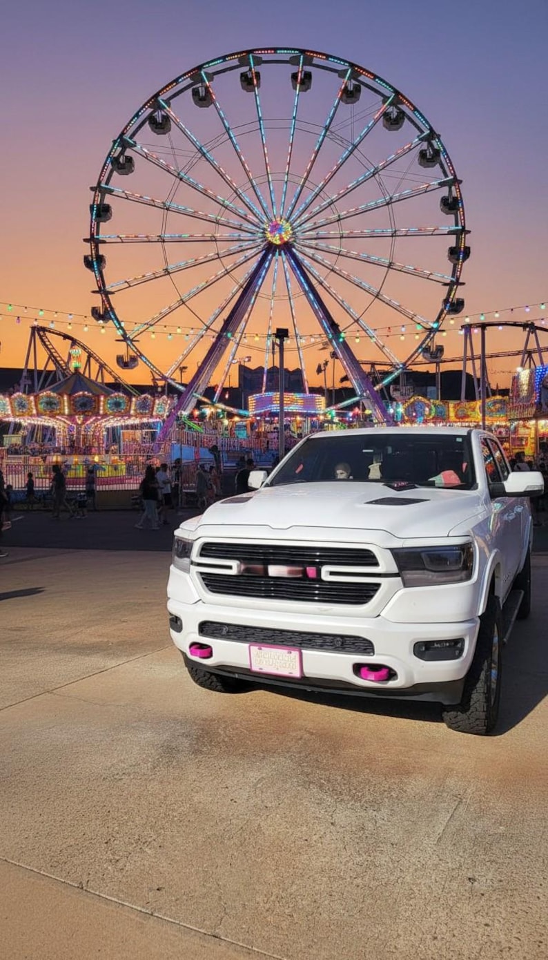 May include: A white pickup truck with pink accents is parked in front of a brightly lit Ferris wheel and carnival rides at dusk. The sky transitions from orange to purple. The truck has a pink license plate.