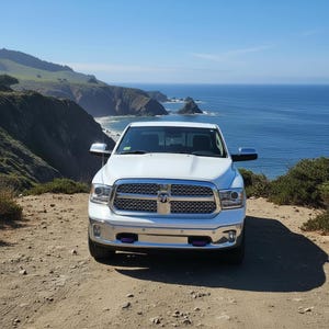 May include: A white Ram pickup truck parked on a dirt road overlooking the ocean. The truck has a chrome grill and accents, with purple tow hooks. The background features cliffs, green hills, and a blue ocean under a clear sky.