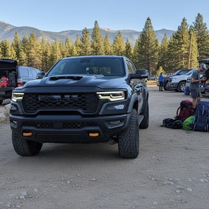 May include: A black pickup truck with a prominent front grille and orange accent lights is parked in a dirt lot. Other vehicles and people are visible in the background, with backpacks and gear suggesting an outdoor activity. Mountains and trees are in the distance.