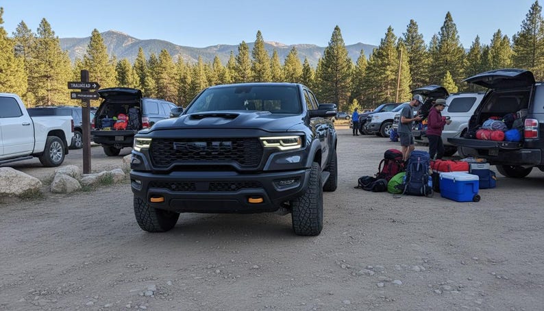 May include: A black pickup truck with a prominent front grille and orange accent lights is parked in a dirt lot. Other vehicles and people are visible in the background, with backpacks and gear suggesting an outdoor activity. Mountains and trees are in the distance.