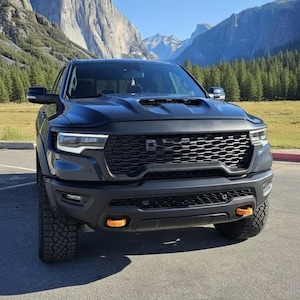 May include: A black pickup truck with a prominent front grille and orange accent lights. The truck is parked on a paved surface with mountains and a blue sky in the background. The tires are large and rugged.