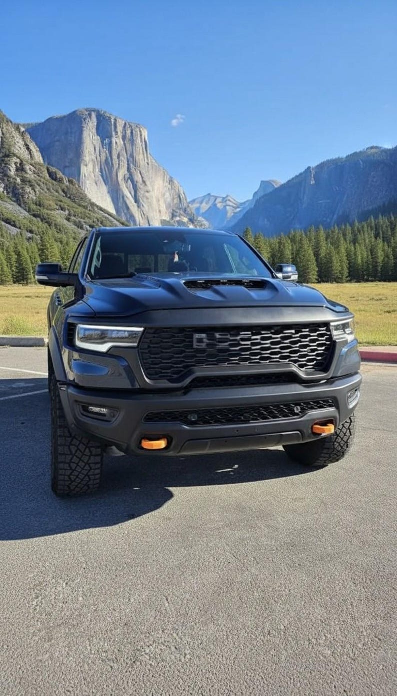 May include: A black pickup truck with a prominent front grille and orange accent lights. The truck is parked on a paved surface with mountains and a blue sky in the background. The tires are large and rugged.