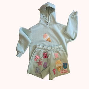 May include: A pale green hoodie and shorts co-ord set. The hoodie and shorts feature an ice cream cone design with glitter detailing. The shorts have colourful letter patches spelling out words and phrases.