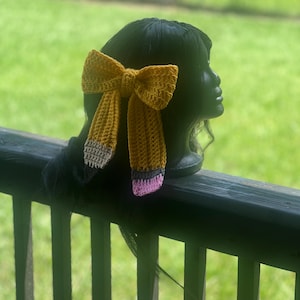 May include: A mustard yellow crochet hair bow, designed to resemble a pencil. The bow features grey, white, and pink details. The hair accessory is displayed on a black wig head, with a green, out-of-focus background.