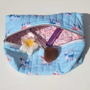 Puede incluir: Una bolsa de maquillaje acolchada de color azul claro con cierre de cremallera. La bolsa está decorada con un estampado de caniche blanco y tiene un forro floral rosa. La bolsa está abierta y contiene un pincel de maquillaje morado y una flor blanca.