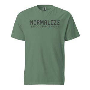 Può includere: T-shirt verde salvia con la scritta "NORMALIZE ENCOURAGEMENT" in grigio scuro. La maglietta a maniche corte e girocollo ha una vestibilità rilassata, adatta per l'uso quotidiano. Il testo è centrato sul davanti.