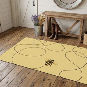 May include: A rectangular area rug with a light yellow background featuring a single honeybee illustration and thin black continuous line patterns that loop across the surface. The rug is positioned on a wooden floor in a room with a wooden bench.