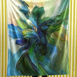 Könnte beinhalten: Eine große Decke mit einem abstrakten Blumendesign. Die Decke hat eine lebendige Farbpalette aus Blau-, Grün- und Gelbtönen, die einen malerischen Effekt erzeugt. Das Design ähnelt Pinselstrichen und verleiht dem Stoff Textur und Tiefe. Die Decke wird von einer Person gehalten.