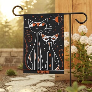 May include: Black garden flag featuring two stylized cats with orange eyes and accents. The word "WELCOME" is printed in orange at the bottom. The flag is displayed on a black metal garden stake.