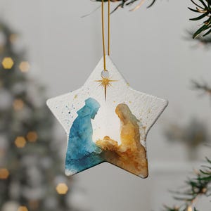 May include: A star-shaped ceramic ornament featuring a watercolor-style nativity scene. The design depicts two figures in blue and gold tones leaning over a central infant, with a gold star positioned above them, all suspended by a gold string.