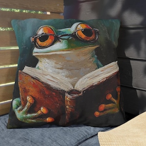 Puede incluir: Cojín decorativo cuadrado con una rana caprichosa con gafas leyendo un libro. La rana está pintada en tonos verdes, naranjas y blancos, con un fondo verde azulado oscuro. El libro es marrón con páginas blancas.