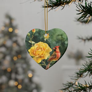May include: Heart-shaped ceramic ornament featuring a watercolor painting of a yellow rose, a red cardinal bird, and green foliage. The ornament is suspended by a gold string, perfect for holiday or seasonal decor.