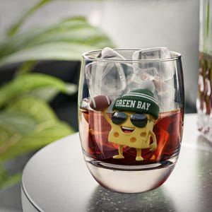 May include: A clear glass tumbler filled with ice cubes and a dark amber liquid. A yellow cheese wedge figurine wearing sunglasses and a green beanie with "GREEN BAY" printed on it is inside the glass, holding a football.