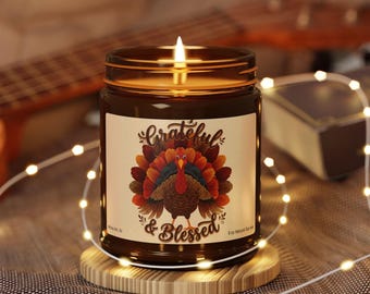 May include: A brown glass jar candle with a lit wick and a label featuring a colorful turkey illustration and the text Grateful & Blessed. The label also includes the text 9 oz Natural Soy wax. The candle sits on a round wooden base.