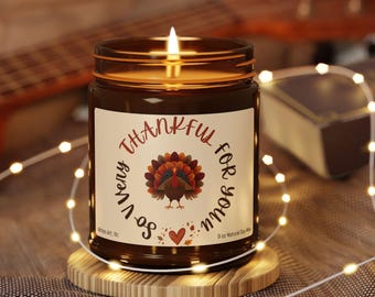 May include: A lit candle in an amber glass jar with a white label featuring a turkey illustration and the text So Very Thankful For You. The label includes a small heart and leaf graphic, and states 9 oz Natural Soy Wax.