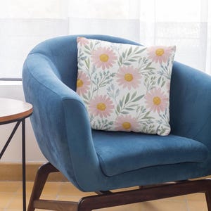 May include: A square decorative pillow with a floral pattern featuring pink daisies with yellow centers and green leaves. The pillow is on a blue armchair, creating a cozy and inviting aesthetic.