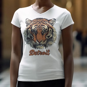 May include: White v-neck t-shirt featuring a detailed tiger face graphic in shades of orange, brown, and black. The word "Detroit" is printed in an orange, retro font below the tiger. The shirt is a casual wear item.