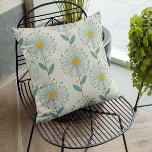May include: A square decorative pillow with a cream background and a repeating pattern of stylized dandelion flowers. The flowers have yellow centers, light blue petals, and green leaves. The pillow is on a black metal chair.