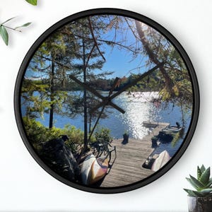 Puede incluir: Un reloj redondo negro con una imagen de una escena de lago. El lago está rodeado de árboles y hay un muelle de madera con dos canoas. La esfera del reloj es negra con números y manecillas blancas.
