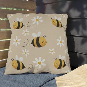 May include: Square beige pillow with a repeating pattern of cartoon bees and white daisies. The bees are yellow and black with white wings, and the daisies have white petals and yellow centers. The pillow is on a blue cushion.