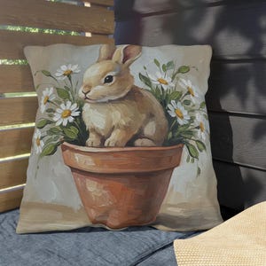 May include: Decorative square pillow featuring a painted image of a brown bunny sitting in a terracotta pot surrounded by white daisies. The pillow has a neutral background and is placed on a blue surface.
