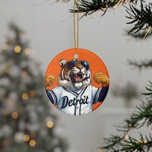 May include: Round orange ceramic ornament featuring a tiger wearing sunglasses and a baseball jersey with "Detroit" on the front. The tiger is flexing its arms. The ornament is hanging from a gold string, perfect for holiday decor.