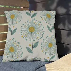 May include: Square throw pillow with a repeating pattern of stylized dandelion flowers. The flowers have yellow centers, teal-colored petals, and teal and yellow accents. The pillow is on a blue surface.