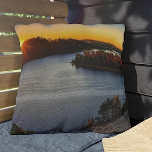 May include: A throw pillow with a scenic sunset over a lake. The pillow features a vibrant orange and yellow sunset with a dark blue lake in the foreground. The lake is surrounded by trees and a small island.