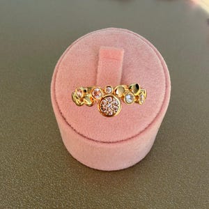 Könnte beinhalten: Ein goldfarbener Ring mit einer Ansammlung kleiner, runder Edelsteine und einem größeren, zentralen Cluster kleinerer Steine. Der Ring wird auf einem rosa Samtringhalter mit rosa Blütenblättern im Hintergrund präsentiert.