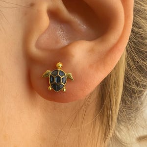 Op de afbeelding: Een close-up van een goudkleurige oorknop in de vorm van een schildpad met een donkerblauwe schild, gedragen op een oor. De oorbel heeft kleine vleugels en staat tegen een lichte achtergrond. Een zeeschelp is wazig op de achtergrond.